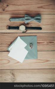Groom accessories for wedding day - watch, textile grey bow tie, rings, grey envelope, white card with copy space, on wooden background, top view