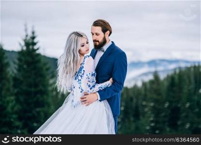 groom in a blue suit and bride in white, embroidered with blue pattern, dress on a background of green pine forests in the mountains of the Carpathians
