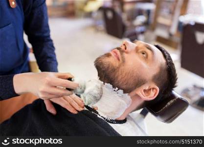 grooming and people concept - man and barber with brush applying shaving foam to beard at barbershop. man and barber applying shaving foam to beard