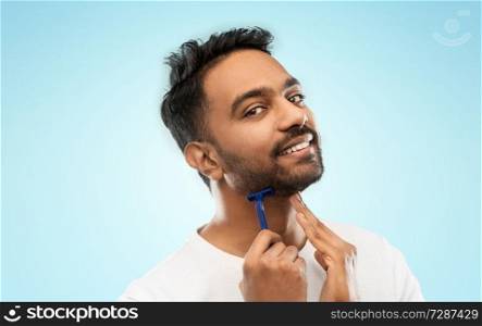 grooming and people concept - smiling indian man shaving beard with manual razor blade over blue background. indian man shaving beard with razor blade
