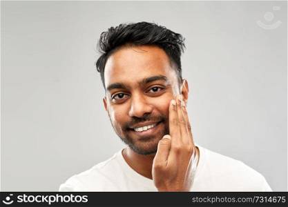 grooming and people concept - smiling indian man touching his beard or applying aftershave over grey background. smiling indian man touching his beard