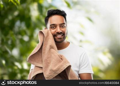 grooming and people concept - smiling indian man with bath towel drying his face over green natural background. indian man with towel over natural background