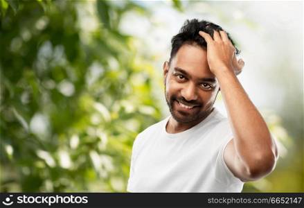 grooming and people concept - smiling young indian man touching his hair over green natural background. indian man touching his hair