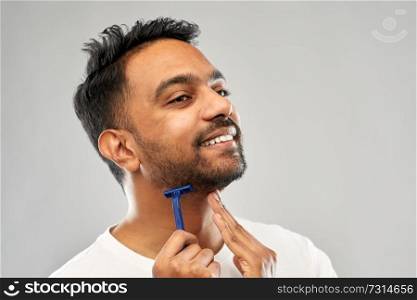 grooming and people concept - young indian man shaving beard with manual razor blade over grey background. indian man shaving beard with razor blade