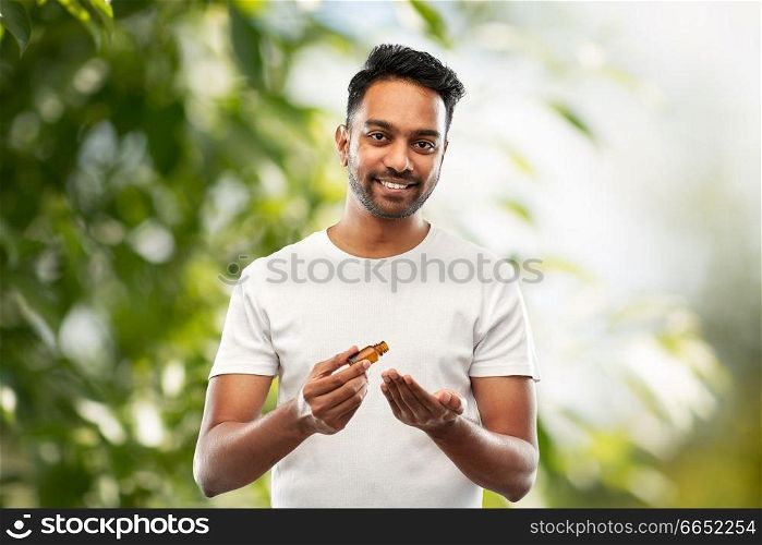 grooming, aromatherapy and people concept - smiling young indian man applying essential oil to his hand over green natural background. indian man applying grooming oil to his hand