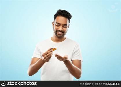 grooming, aromatherapy and people concept - smiling young indian man applying essential oil to his hand over blue background. indian man applying grooming oil to his hand