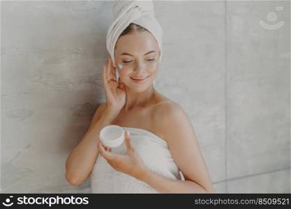 Grooming, cosmetology and wellness concept. Pleased female model poses with jar of moisturizing cream, applies cosmetic product on cheek, enjoys self care poses wrapped in towel near grey wall