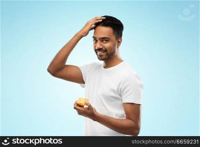grooming, hairstyling and people concept - smiling young indian man applying hair wax or styling gel over blue background. indian man applying hair wax or styling gel