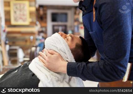 grooming, shaving and people concept - barber softening male face skin with hot towel at barbershop. barber softening male face sking with hot towel