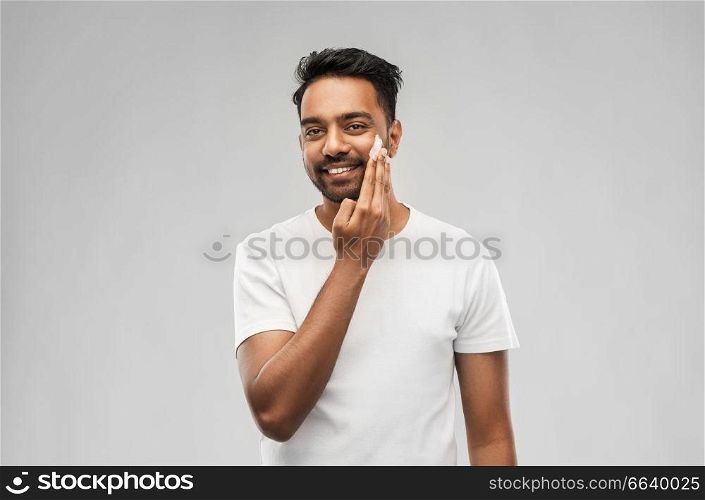 grooming, skin care and people concept - smiling young indian man applying cream to face over gray background. happy indian man applying cream to face