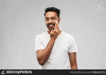 grooming, skin care and people concept - smiling young indian man applying cream to face over gray background. happy indian man applying cream to face