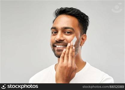 grooming, skin care and people concept - smiling young indian man applying cream to face over grey background. happy indian man applying cream to face