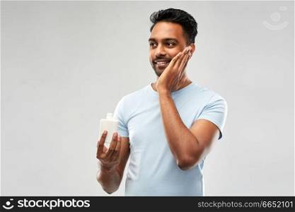 grooming, skin care and people concept - smiling young indian man applying lotion to face over grey background. happy indian man applying lotion to face