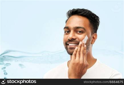 grooming, skin care and people concept - smiling young indian man applying cream to face over blue background with bubbles in water splash. happy indian man applying cream to face