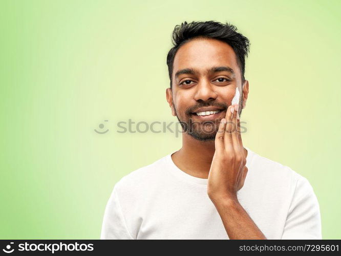 grooming, skin care and people concept - smiling young indian man applying cream to face over lime green natural background. happy indian man applying cream to face