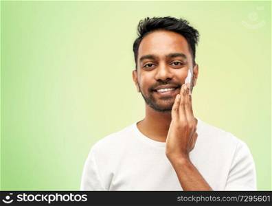 grooming, skin care and people concept - smiling young indian man applying cream to face over lime green natural background. happy indian man applying cream to face