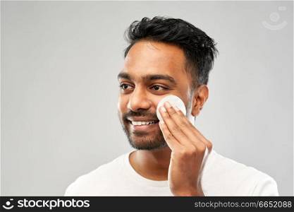 grooming, skin care and people concept - smiling young indian man cleaning his face with cotton pad over grey background. smiling indian man cleaning face with cotton pad