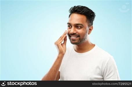 grooming, skin care and people concept - smiling young indian man cleaning his face with cotton pad over blue background. smiling indian man cleaning face with cotton pad