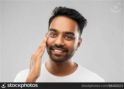 grooming, skin care and people concept - smiling young indian man touching his face over grey background. smiling indian man touching his face