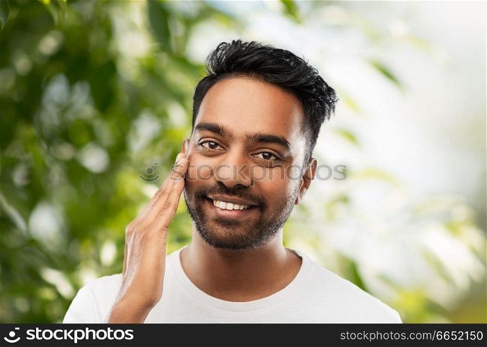 grooming, skin care and people concept - smiling young indian man touching his face over green natural background. smiling indian man touching his face