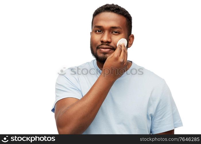 grooming, skin care and people concept - young african american man cleaning his face with cotton pad over white background. african american man cleaning face with cotton pad