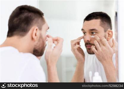 grooming, skin care and people concept - young man applying cream to face and looking to mirror at home bathroom. young man applying cream to face at bathroom
