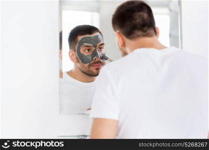 grooming, skin care and people concept - young man with brush applying clay mask at home bathroom mirror. young man applying clay mask to face at bathroom