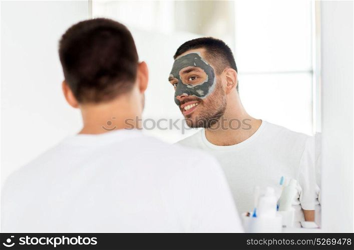 grooming, skin care and people concept - young man with clay mask on face and looking to mirror at home bathroom. young man with clay mask on face at bathroom