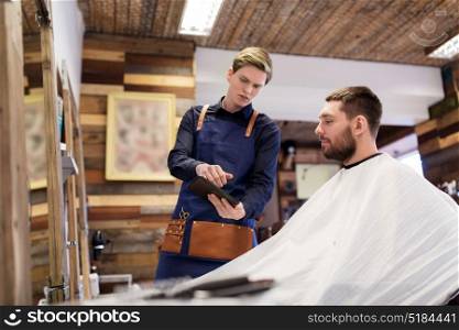 grooming, technology and people concept - hairdresser or barber showing tablet pc computer to man at barbershop. barber showing tablet pc to man at barbershop