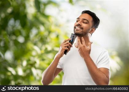 grooming, technology and people concept - smiling indian man shaving beard with trimmer over green natural background. smiling indian man shaving beard with trimmer