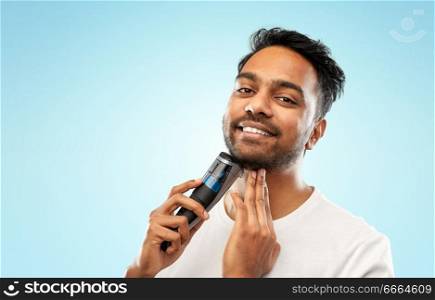 grooming, technology and people concept - smiling indian man shaving beard with trimmer over blue background. smiling indian man shaving beard with trimmer
