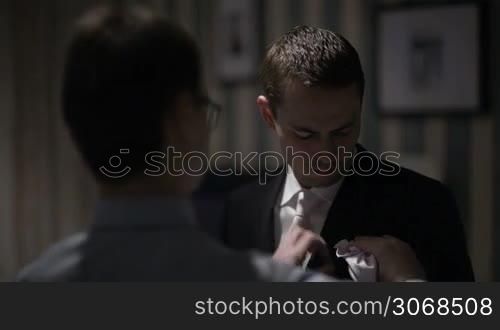 Groomsman putting white handkerchief into pocket of groom&acute;s jacket. Preparation before wedding ceremony.