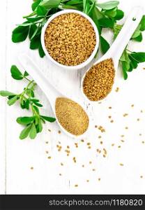 Ground fenugreek in a spoon, spice seeds in a bowl and spoon with green leaves on wooden board background from above
