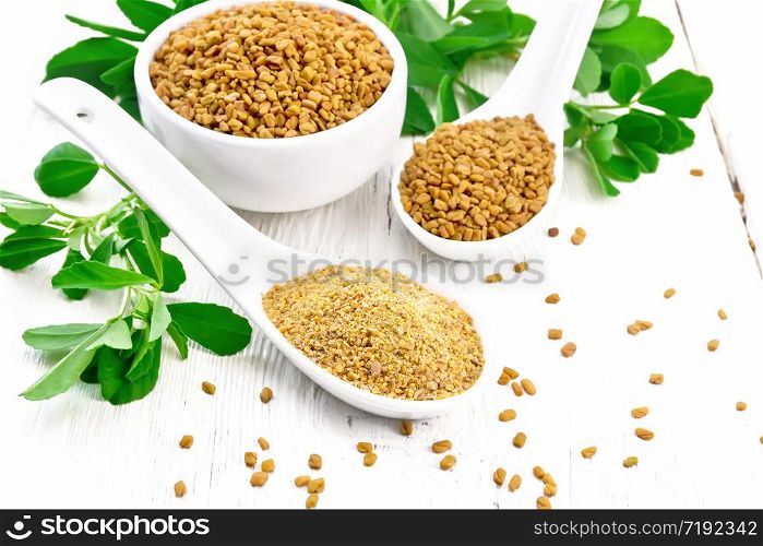 Ground fenugreek in a spoon, spice seeds in a bowl and spoon with green leaves on white wooden board background