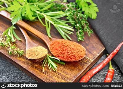 Ground hot red pepper and paprika in spoons, chili pods, spicy greens and a napkin on black wooden board background