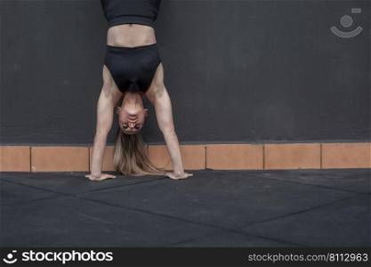 Ground level of muscular sportswoman in activewear doing handstand exercise near black wall during fitness workout in gym. Female athlete doing handstand during training