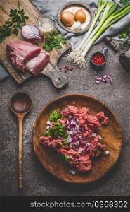 Ground meat in plate with cooking spoon with ingredients on rustic kitchen table background, top view