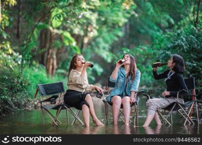Group beautiful Asian women friends trave≤rs relaxing in c&chairs  in stream, They are cheering and drinking beer during c&ing, talking with fun and happy to≥ther