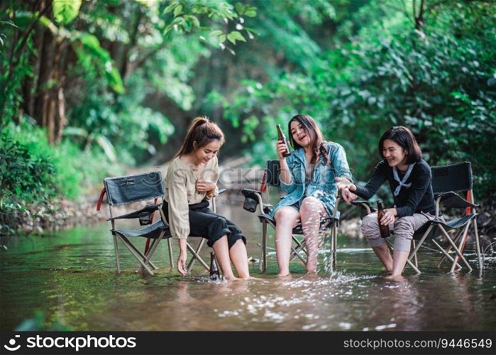 Group beautiful Asian women friends travelers relaxing in c&chairs  in stream, They are cheering and drinking beer during c&ing, talking with fun and happy together