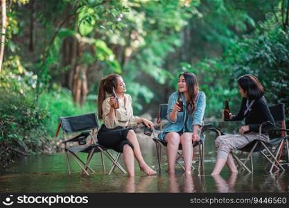 Group beautiful Asian women friends travelers relaxing in c&chairs  in stream, They are cheering and drinking beer during c&ing, talking with fun and happy together