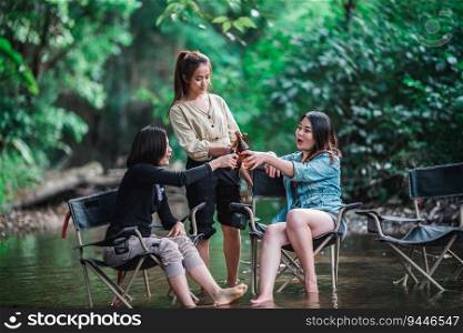 Group beautiful Asian women friends travelers relaxing in camp chairs  in stream, They are cheering and drinking beer during camping, talking with fun and happy together