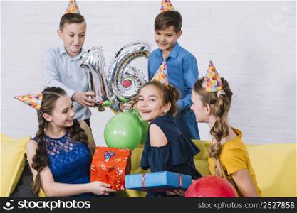 group friends celebrating birthday by giving presents holding sliver number 16 foil balloon