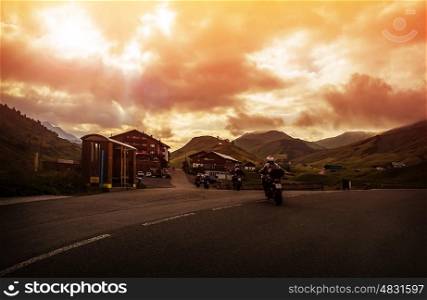 Group of bikers travel across mountainous village, red cloudy sly, overcast weather, Europe, Austria, Alps, active lifestyle, freedom concept