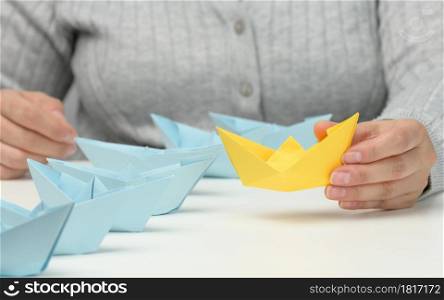 group of blue paper boats follows a yellow boat in front of a white table. The concept of a strong and charismatic leader in a team, manipulating the masses