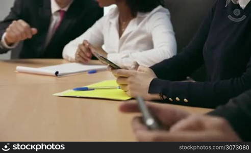 Group of business people during meeting in corporate conference room. A woman types messages and emails on mobile phone. Closeup of hands