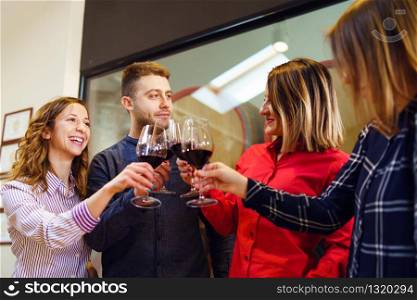 Group of caucasian friends or family standing by the table at winery or restaurant holding a glasses of red wine toasting celebrating young man and three women wearing shirt smiling