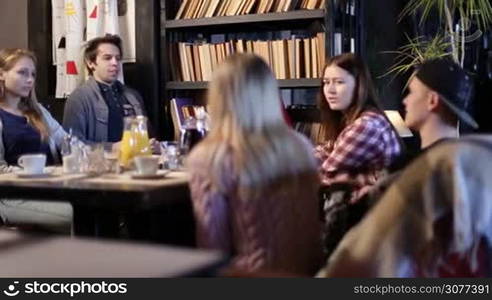 Group of cheerful teenage friends sitting at the table and talking during lunch in art cafe while waitress serving coffee. Smiling classmates relaxing after studying at the restaurant.