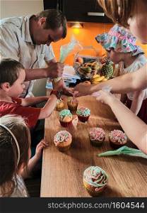 Group of children baking cupcakes, squeezing cream from confectionery bag, preparing ingredients, topping, sprinkles for decorating cookies. Kids cooking, working together in kitchen at home. Concept of happy family