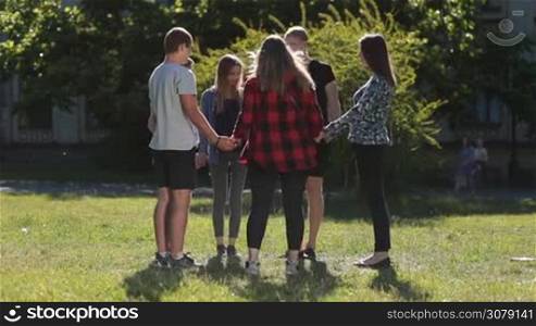Group of christian college students in circle holding hands and praying for good exam results while standing on green grass on university campus. Young teenage friends showing unity on park lawn.