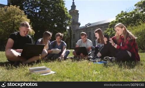 Group of college students working with laptops and digital tablet pc on university campus while sitting on green grass on a sunny day. Dolly shot. Positive teenage friends using modern technology devices while doing homework in the park.